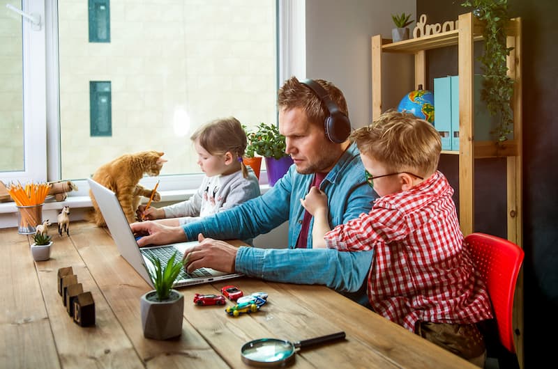 Ein Mann sitzt mit Kindern und Katze am Schreibtisch, ein Beispiel für Work-Life-Blending