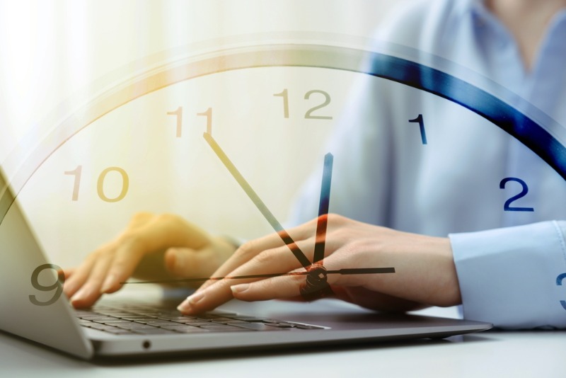 Eine Frau tippt auf Laptoptastatur Uhr im Vordergrund, Arbeitszeit bei Zeitumstellung