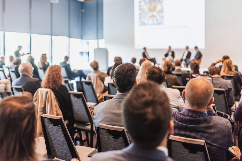 In einem Konferenzraum sitzen Geschäftsleute und hören den Rednern auf der Bühne zu. Sie nehmen an einem Seminar teil und wollen ihre Fachkompetenz verbessern.