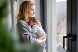 Eine Frau steh am Fenster und hält ihr Baby im Arm. Sie hat ein ruhendes Arbeitsverhältnis während der Elternzeit.