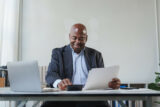 Ein Man im Anzug sitzt im Büro am Schreibtisch. Er hält ein Dokument in der Hand, tippt auf dem Taschenrechner und lächelt dabei. Er freut ich über die Vorteile seiner Entgeltumwandlung.