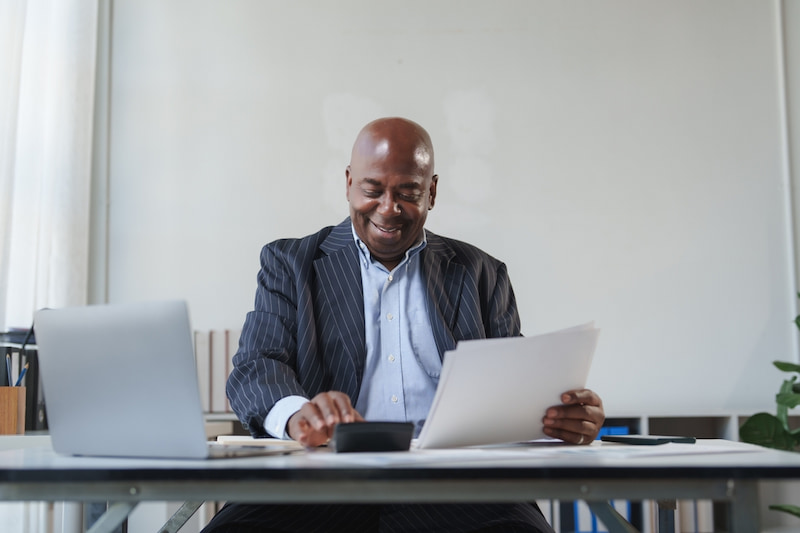 Entgeltumwandlung: Vorteile, Nachteile & Möglichkeiten 2 Ein Man im Anzug sitzt im Büro am Schreibtisch. Er hält ein Dokument in der Hand, tippt auf dem Taschenrechner und lächelt dabei. Er freut ich über die Vorteile seiner Entgeltumwandlung.